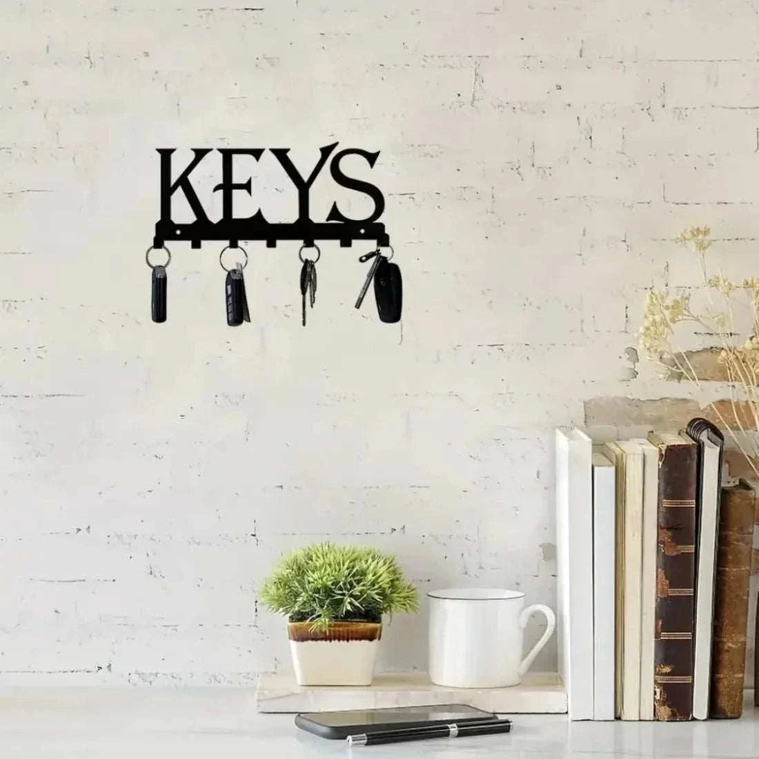 wall mounted black metal key holder with keys hanging on white brick wall above table with books plant and mug