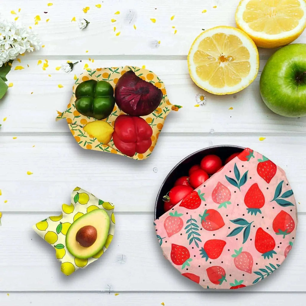 Reusable beeswax food wraps with lemon, strawberry, and fruit patterns covering vegetables on a white wooden surface
