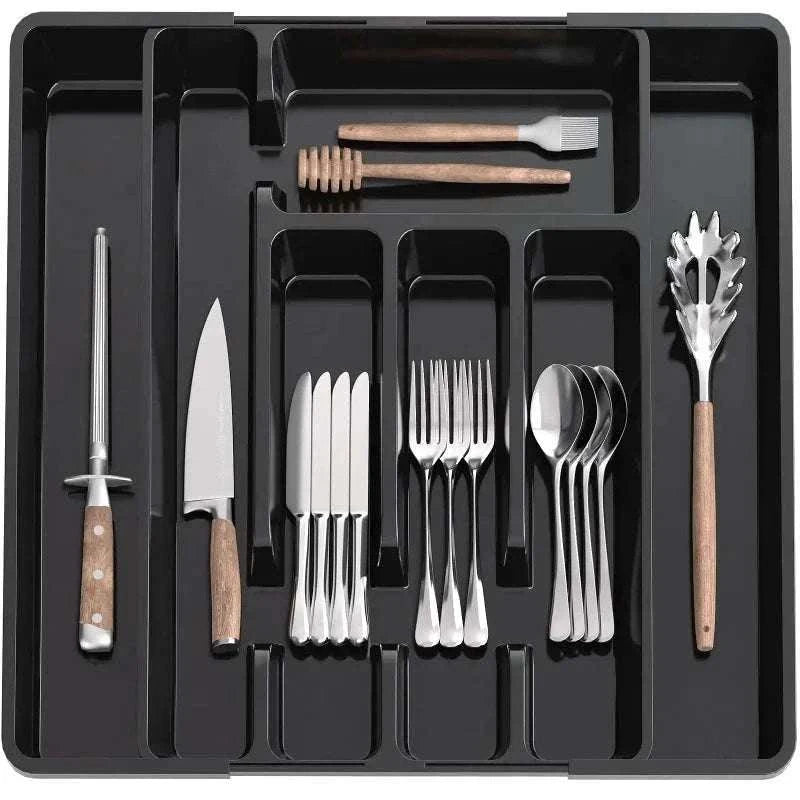 Adjustable black plastic pantry organizer tray with multiple compartments holding silverware and wooden-handled kitchen utensils